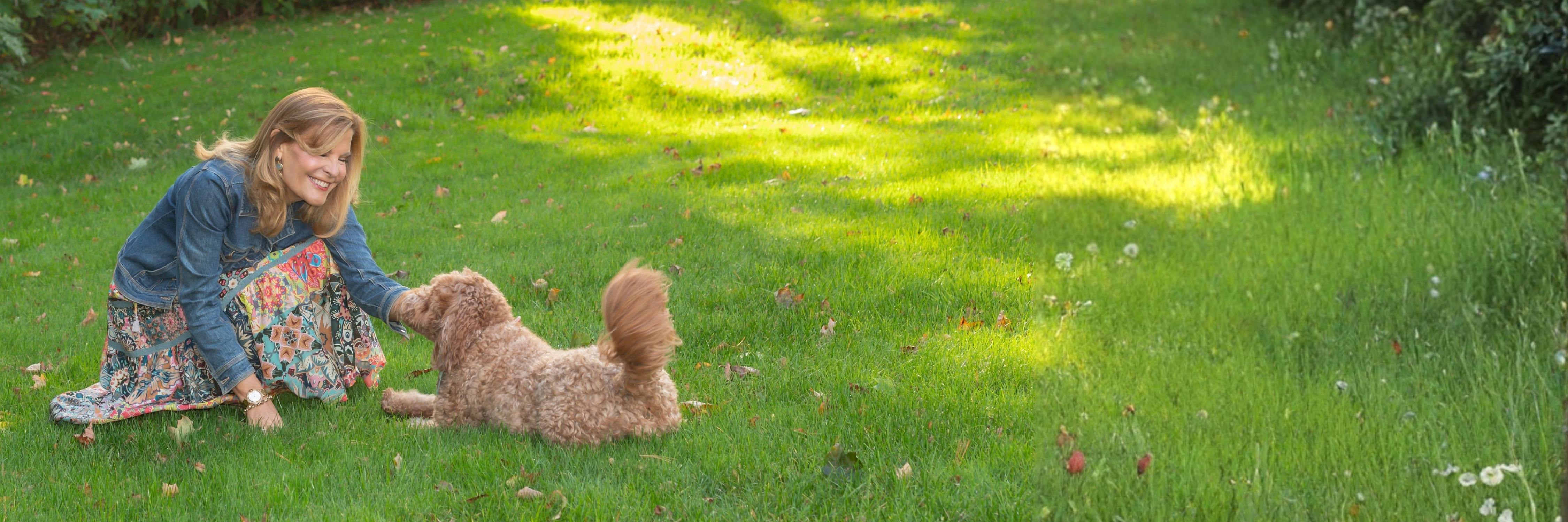 Leighanne kneeling on a sunlit lawn playing with her curly apricot doodle dog