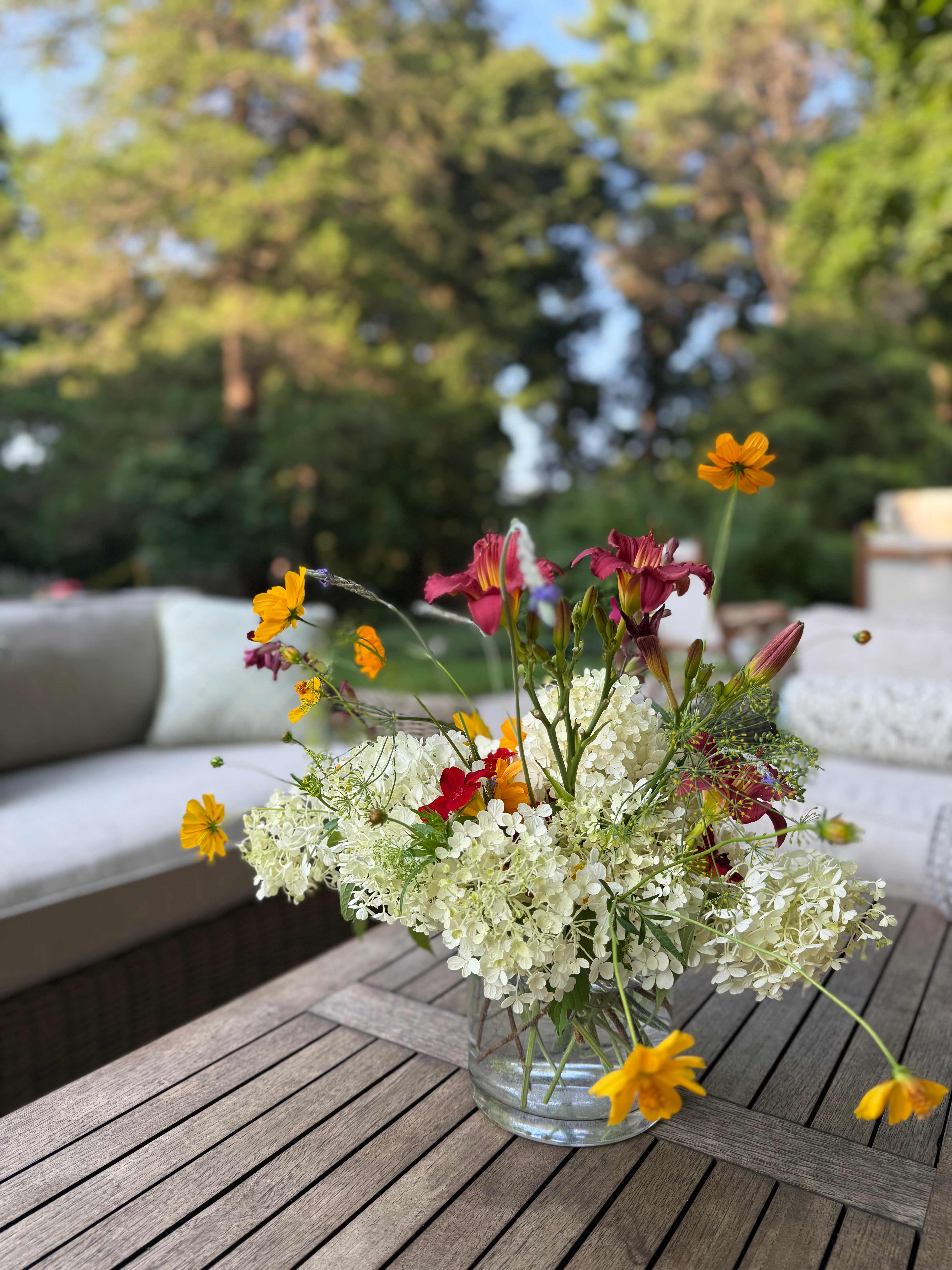 A glass vase of white hydrangeas and orange and red wildflowers on a wooden outdoor table