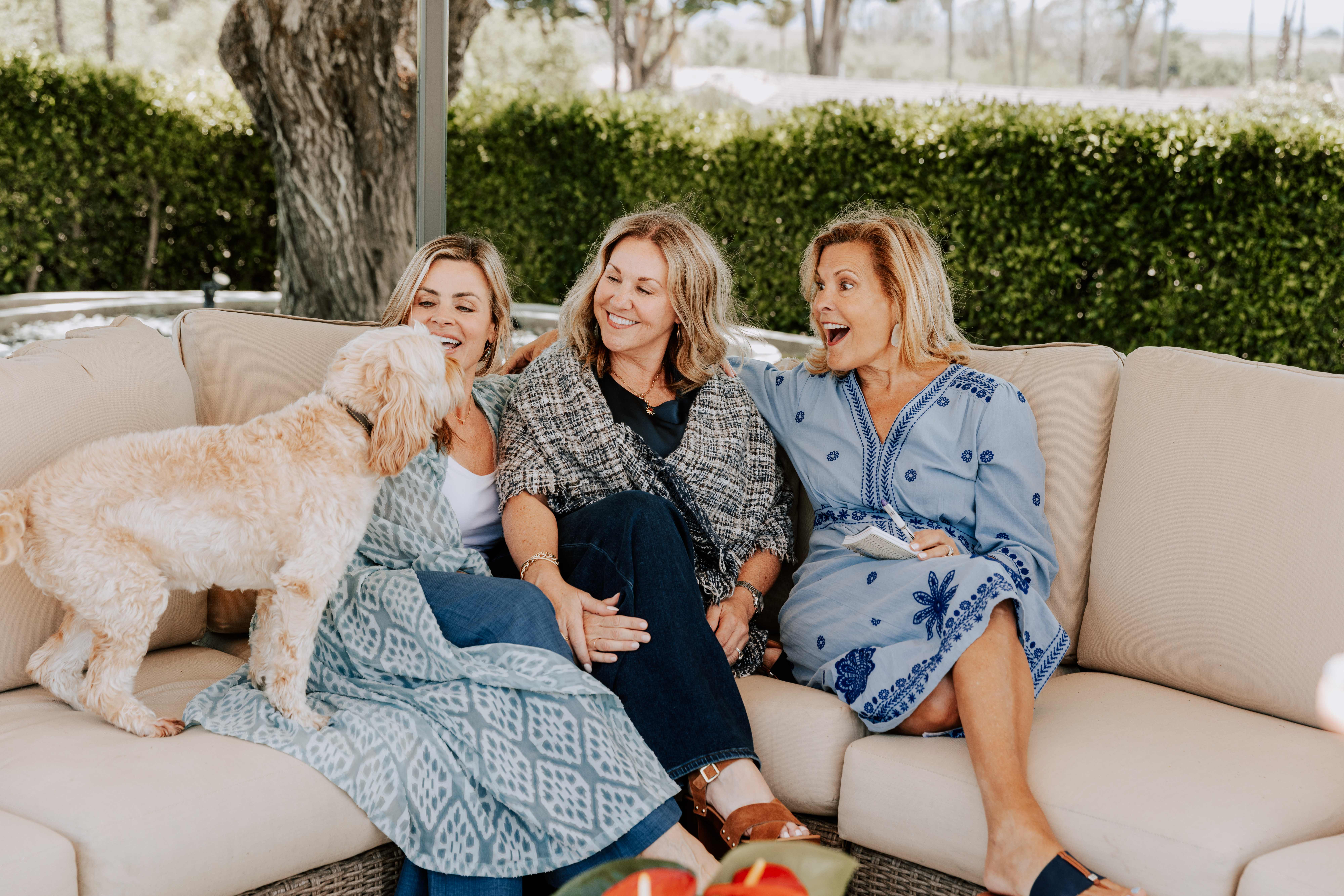 Three women laughing together on a cream outdoor couch with a curly dog beside them
