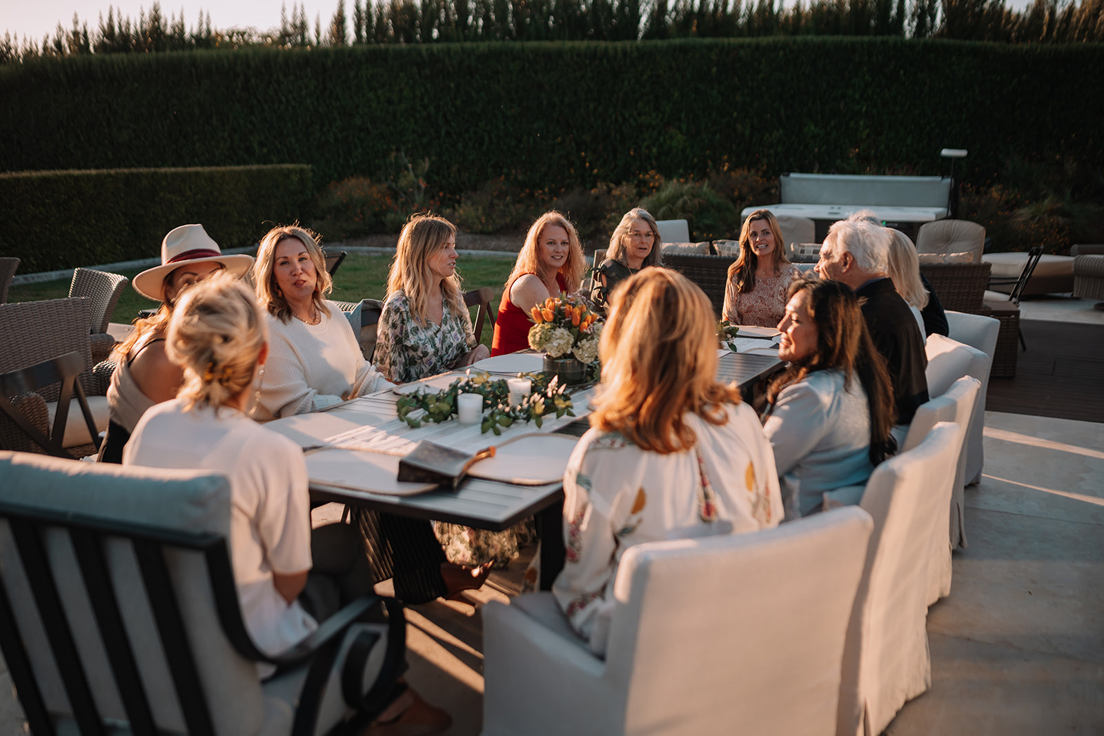 A group seated around a long outdoor dinner table at golden hour with flowers and candles