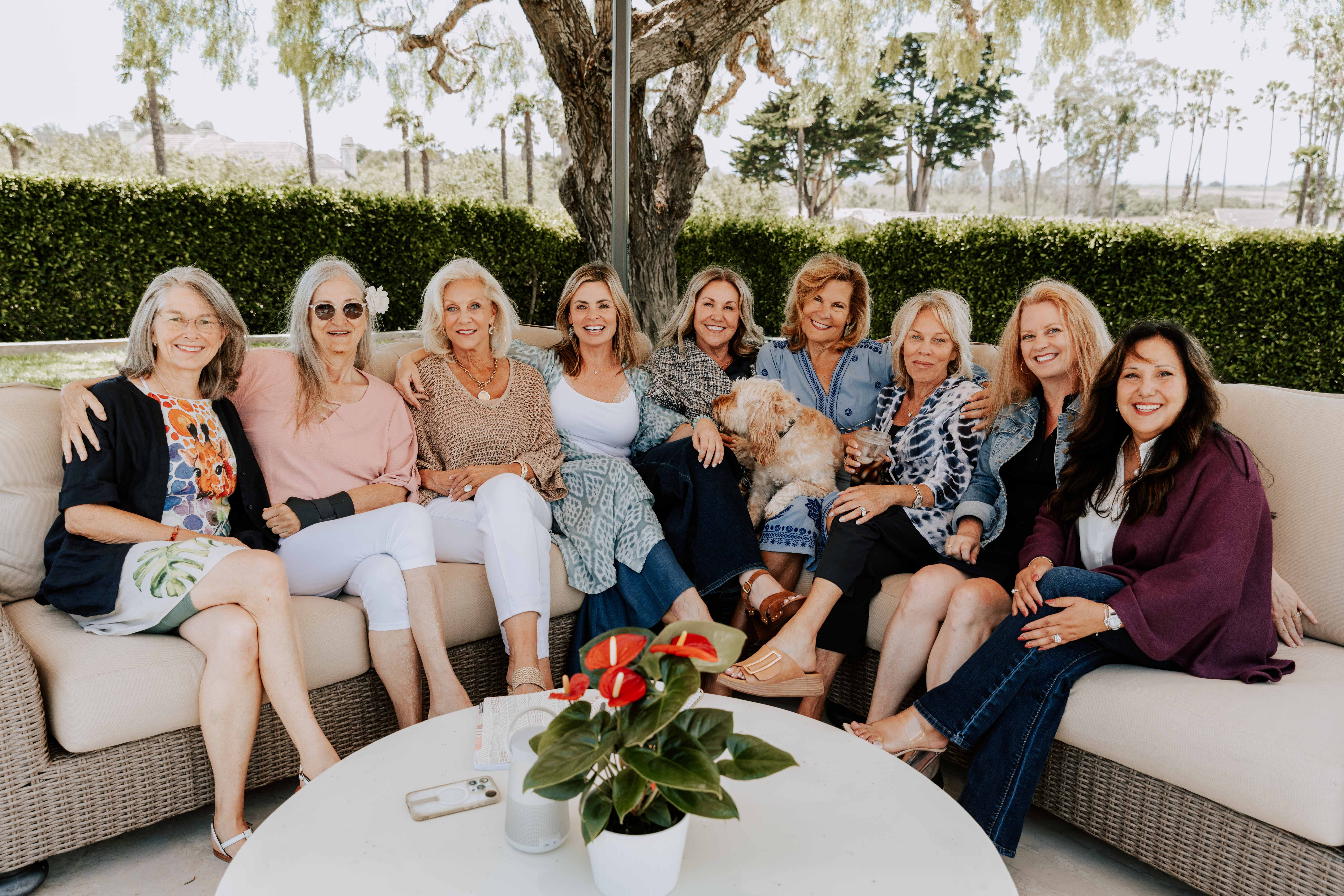 Nine women seated shoulder to shoulder along a curved outdoor couch, smiling, with a dog in the middle